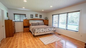 Une chambre avec un plancher en bois, un lit recouvert d’une couvre-lit à motifs, deux fenêtres équipées de stores et un petit tapis.
