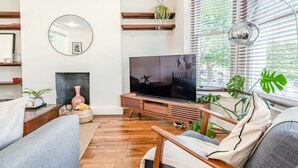 Ett vardagsrum med en TV, trämöbler, en öppen spis och stora fönster med persienner.