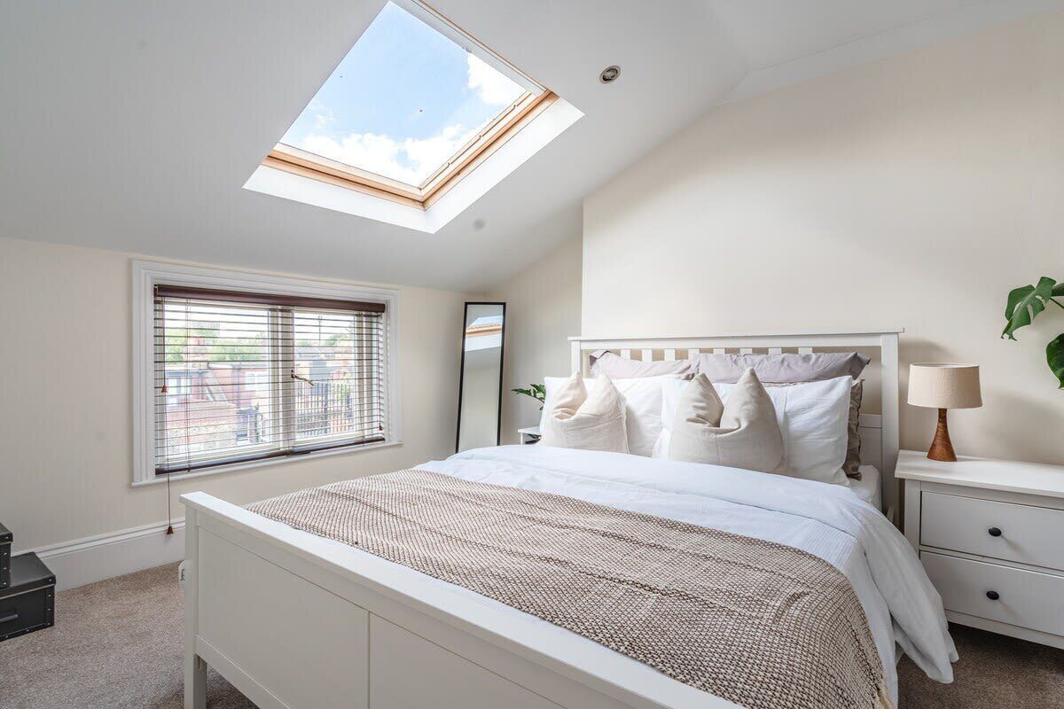 A bedroom with a skylight, a bed with white bedding, a bedside table with a lamp, and a window with blinds.