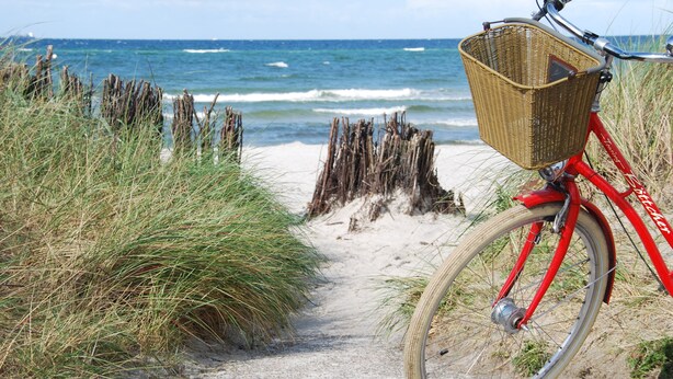 Uma bicicleta vermelha com cesto de vime estacionada em um caminho de areia ao lado do oceano.