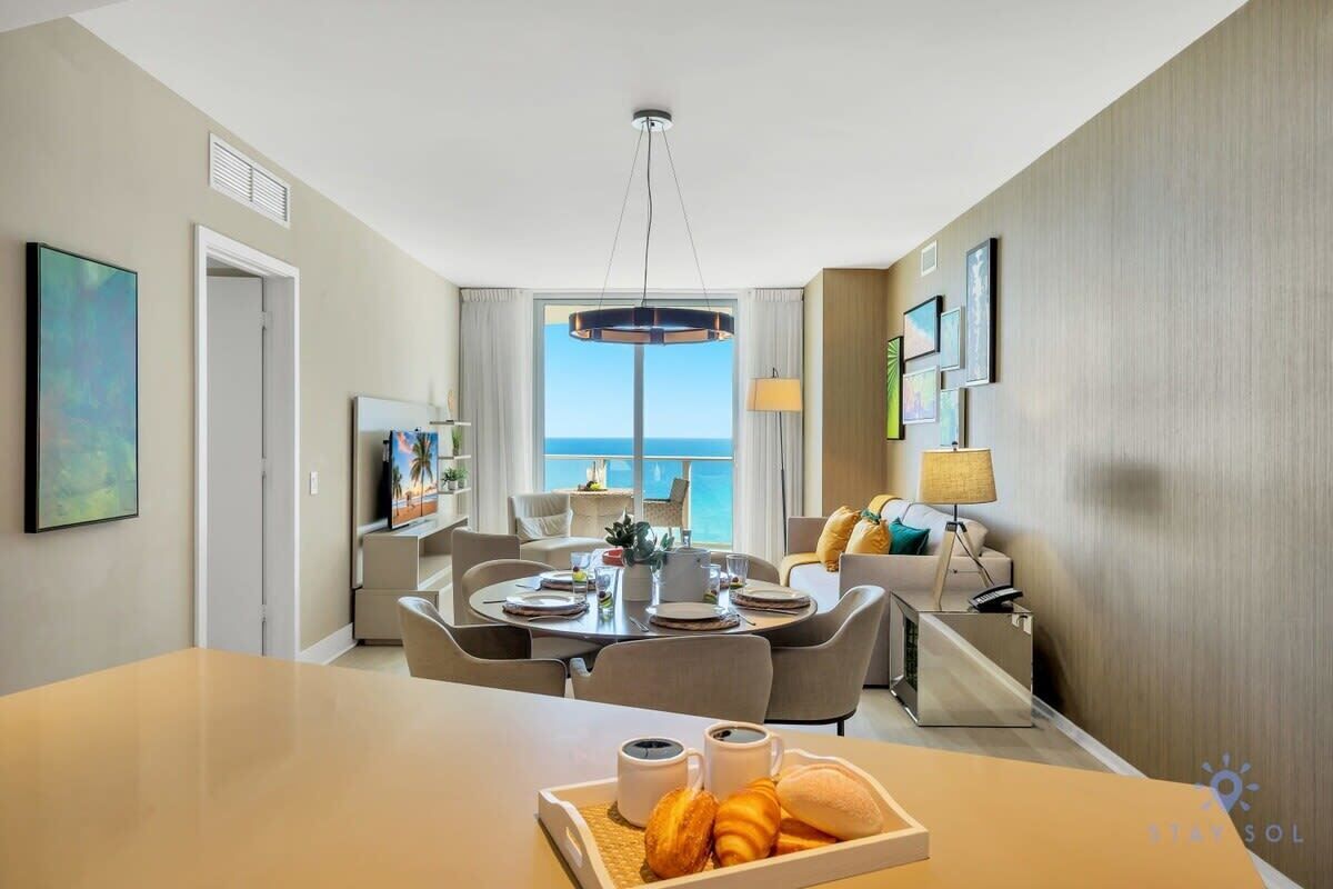 A modern hotel room with a dining area, a large window with a view of the ocean, and a balcony with a view of the sea.