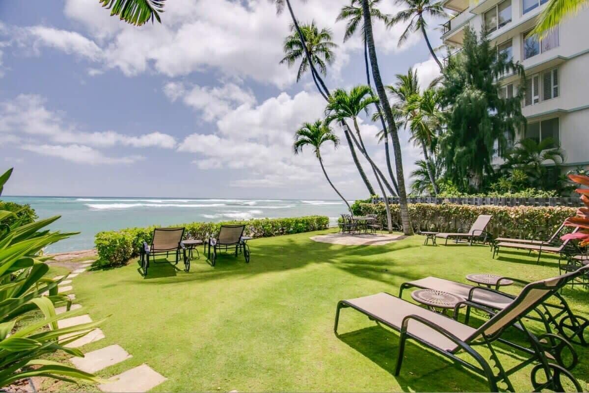 A well-maintained outdoor area with a lawn, lounge chairs, and tables, overlooking a beach with palm trees and a building in the background.