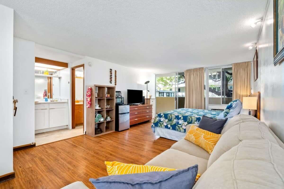 Une chambre spacieuse avec un grand lit, un canapé et une télévision. On aperçoit un coin cuisine à travers une porte ouverte.