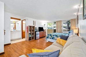 A spacious bedroom with a large bed, a sofa, and a TV. There is a kitchen area visible through an open door.