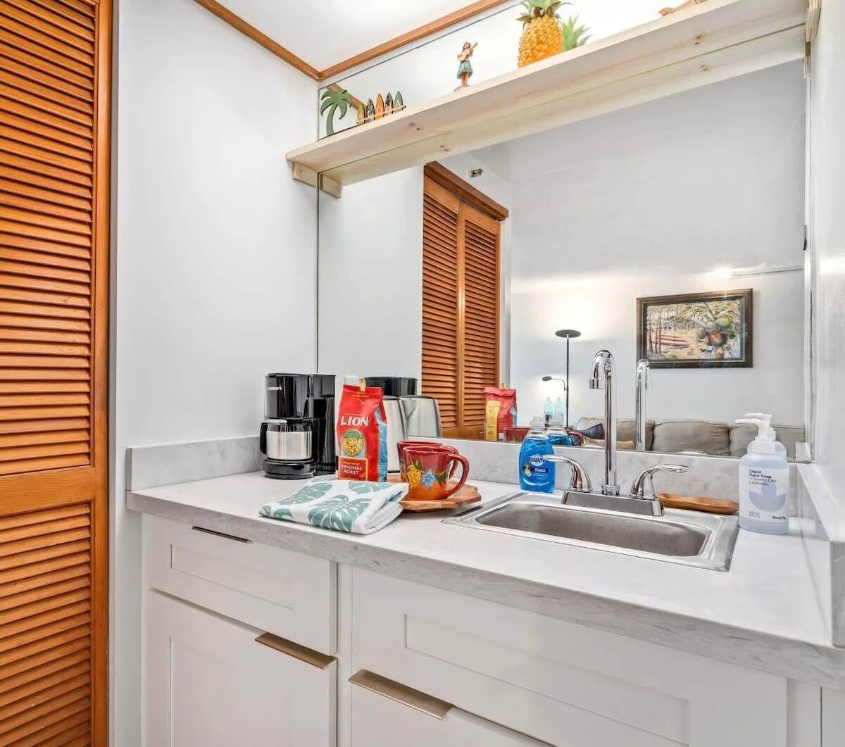 A modern bathroom with a white countertop, a sink, and a mirror. There are toiletries and a coffee maker on the counter.