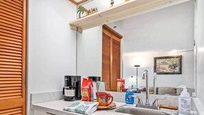 A modern bathroom with a white countertop, a sink, and a mirror. There are toiletries and a coffee maker on the counter.