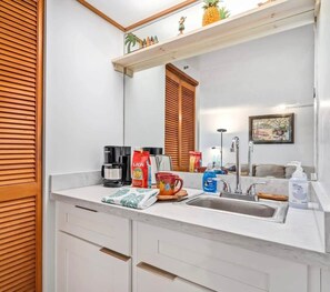 A modern bathroom with a white countertop, a sink, and a mirror. There are toiletries and a coffee maker on the counter.