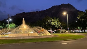 Une fontaine bien éclairée, dotée d’une ouverture centrale, entourée d’une zone pavée bordée d’une pelouse, le tout se dressant sur fond de montagnes.