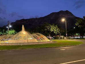 A well-lit fountain with a central spout, surrounded by a paved area with a grassy border, set against a mountain backdrop.