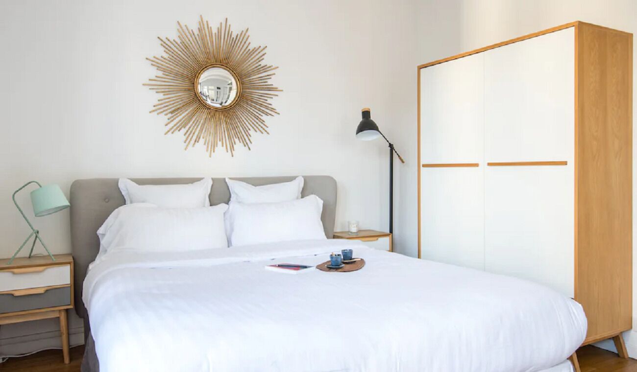 A neatly made bed with white linens, a bedside lamp, a small bedside table with a lamp, and a large wardrobe.