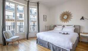 Un dormitorio con una cama grande, una silla, una mesita de noche, un radiador y una ventana con cortinas.