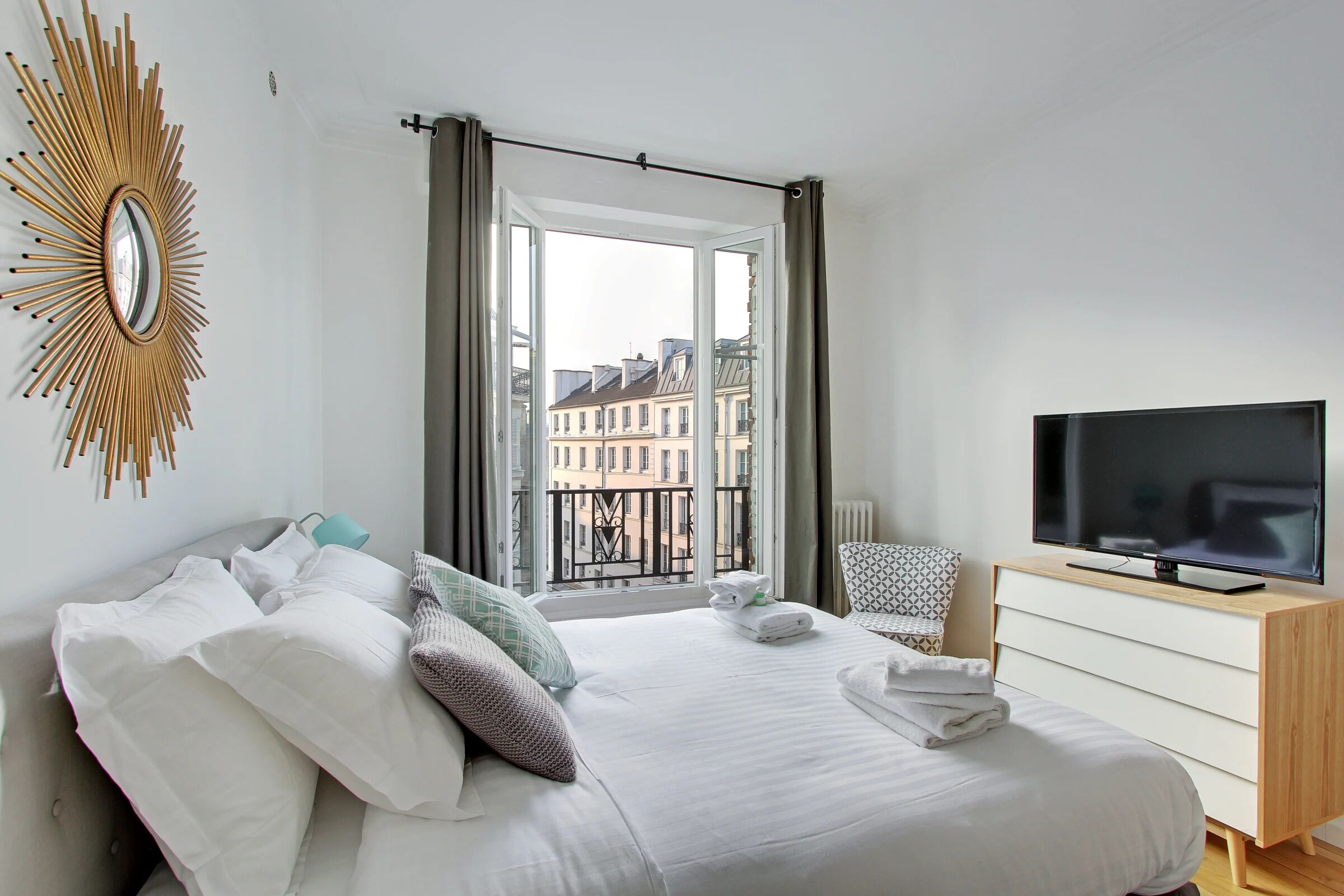 A hotel room with a bed, a TV, a dresser, and a view of buildings through a window.