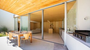 A modern kitchen with a large island, sliding glass doors leading to a dining area, and a patio with a wooden table and chairs. - Villa Maupiti Avec Piscine Chauffée (HOSSEGOR)