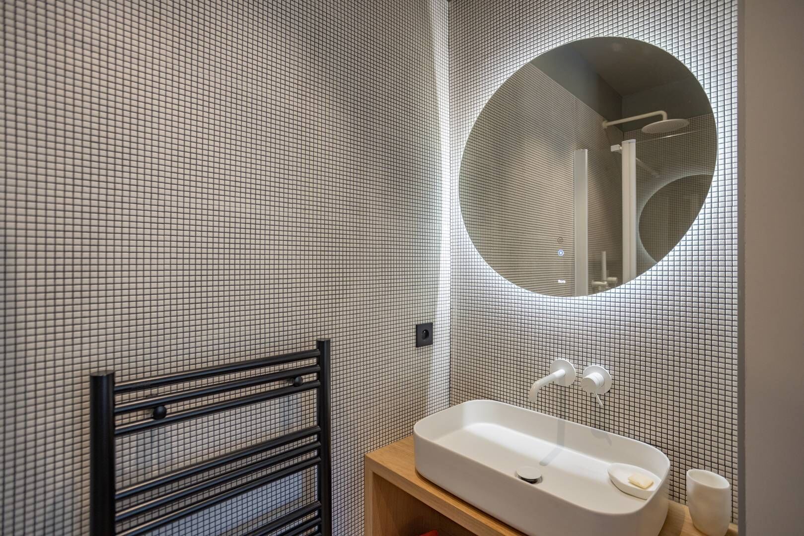 A bathroom with a tiled wall, a round mirror, a white sink, and a black metal rack.