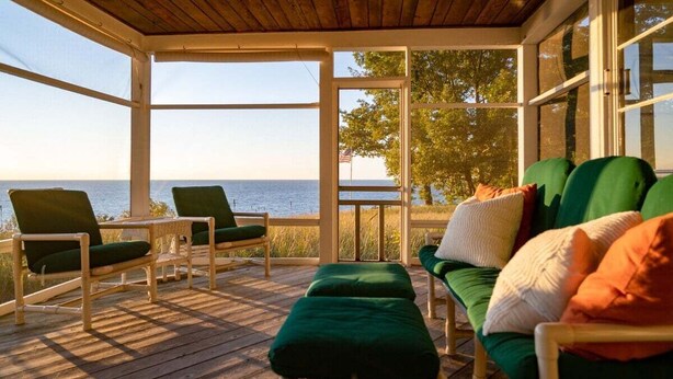 Ett takförsett utomhusområde med sittplatser och utsikt över havet, trägolv och grönt klädd möblemang.