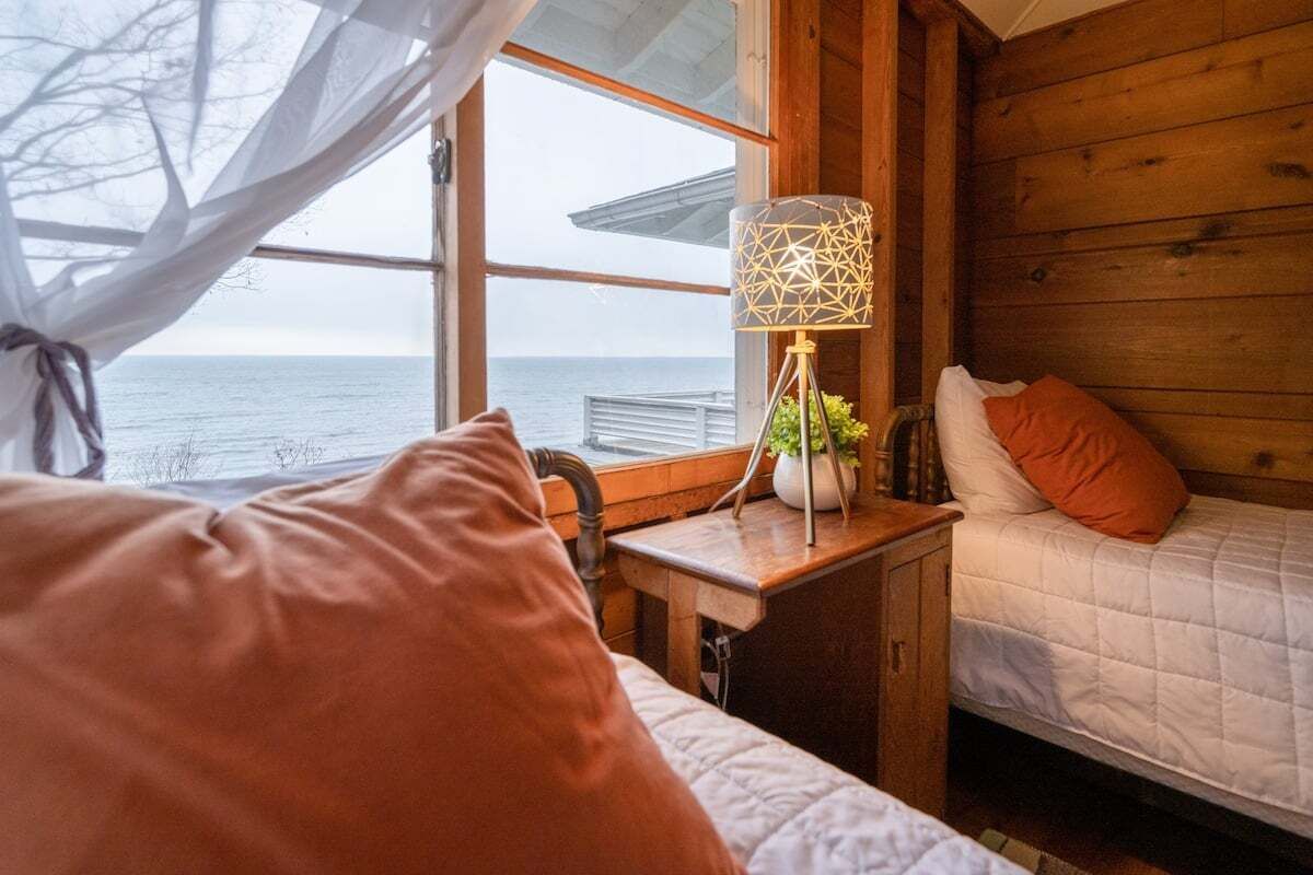 Une chambre confortable dans un chalet, avec un lit, une table de chevet dotée d’une lampe, et une vue sur la mer par la fenêtre.