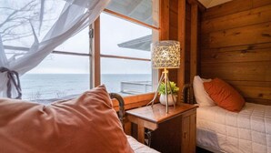 Ett mysigt rum i en stuga med en säng, ett sängbord med en lampa och utsikt över havet genom ett fönster.