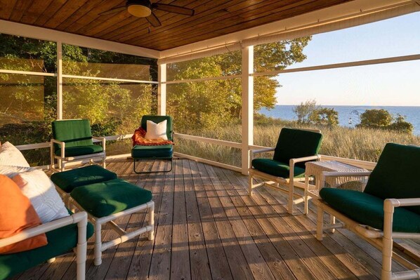 A covered outdoor deck with wooden flooring and white railings, featuring green cushioned chairs and a small table.