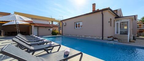 A swimming pool with lounge chairs and umbrellas, a building with a tiled roof, and a clear sky.