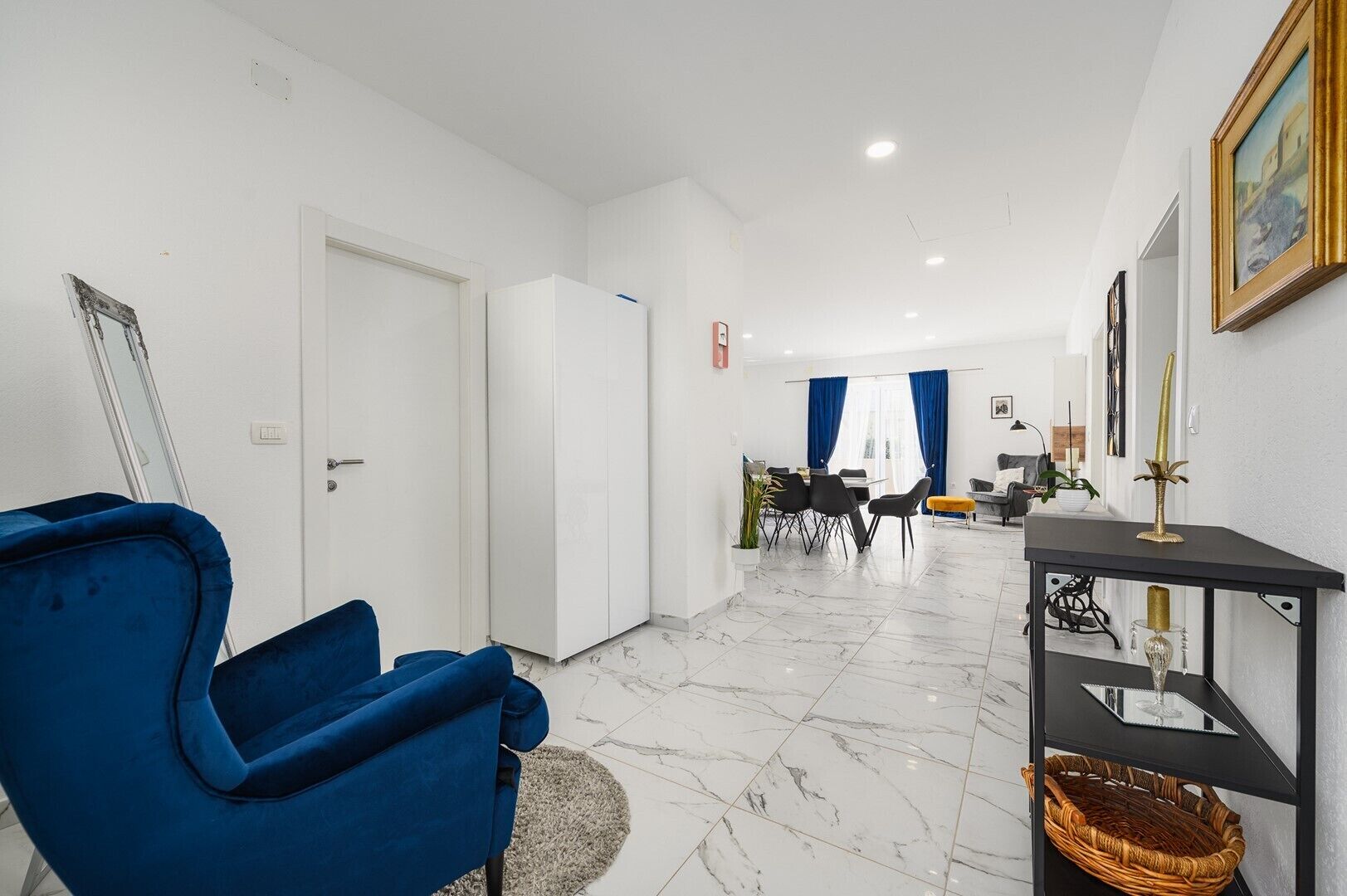 A modern living room with a blue armchair, a dining area with a table and chairs, and a black console table with decorative items.