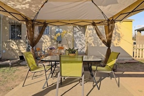 Un patio exterior con mesa y sillas, una zona cubierta con toldo blanco y plantas en macetas.