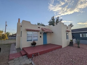 Una casa pequeña con un porche delantero rojo, una puerta azul y un pequeño jardín con grava y plantas.