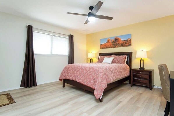 Un dormitorio con cama, dos lámparas de noche, ventilador de techo, ventana con cortinas y un cuadro en la pared.