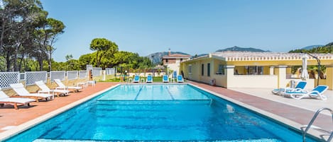 A swimming pool with lounge chairs, a building with a yellow facade, and trees in the background.