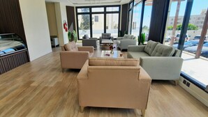 A modern hotel lobby with a wooden floor, beige sofas, a glass partition, and large windows offering a view of the street.