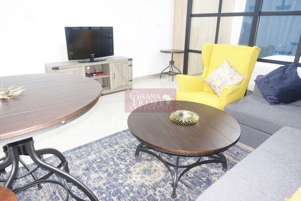A living room with a round wooden table, a yellow armchair, a grey sofa, and a television on a wooden cabinet.