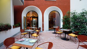 Un área de patio al aire libre con mesas, sillas, entrada de un edificio y plantas en macetas.