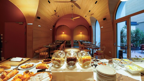 Un bufé de desayuno con diversos alimentos y recipientes de vidrio transparente en un comedor moderno con techo de madera y grandes ventanales.