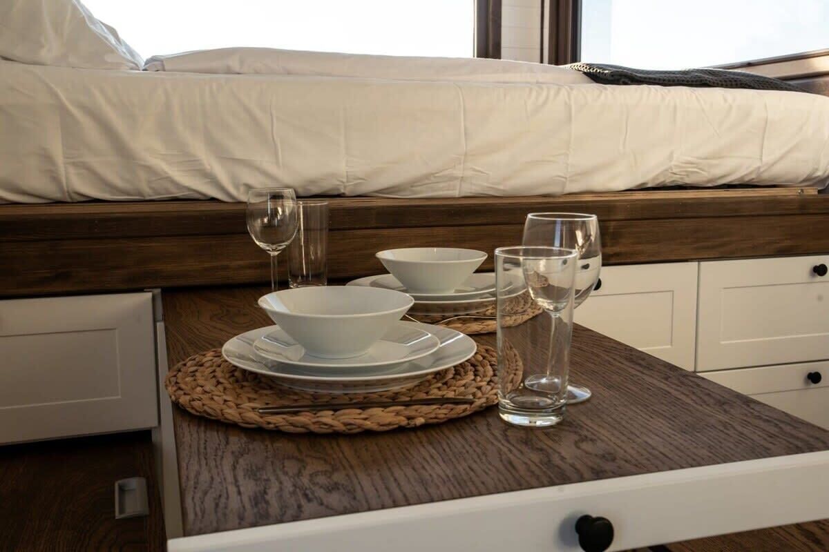 Une table de petit-déjeuner soigneusement dressée, avec de l’assiette, des bols et des verres blancs, posée sur un set de table en fibres tressées, dans une pièce moderne où l’on aperçoit un lit au fond.