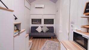 Quarto pequeno com um sofá cinzento, duas janelas, um tapete e uma kitchenette com micro-ondas.