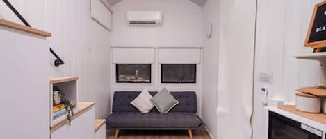 Ein schmales Zimmer mit einer grauen Couch, zwei Fenstern, einem kleinen Teppich und einer Küchenzeile mit einer Mikrowelle.