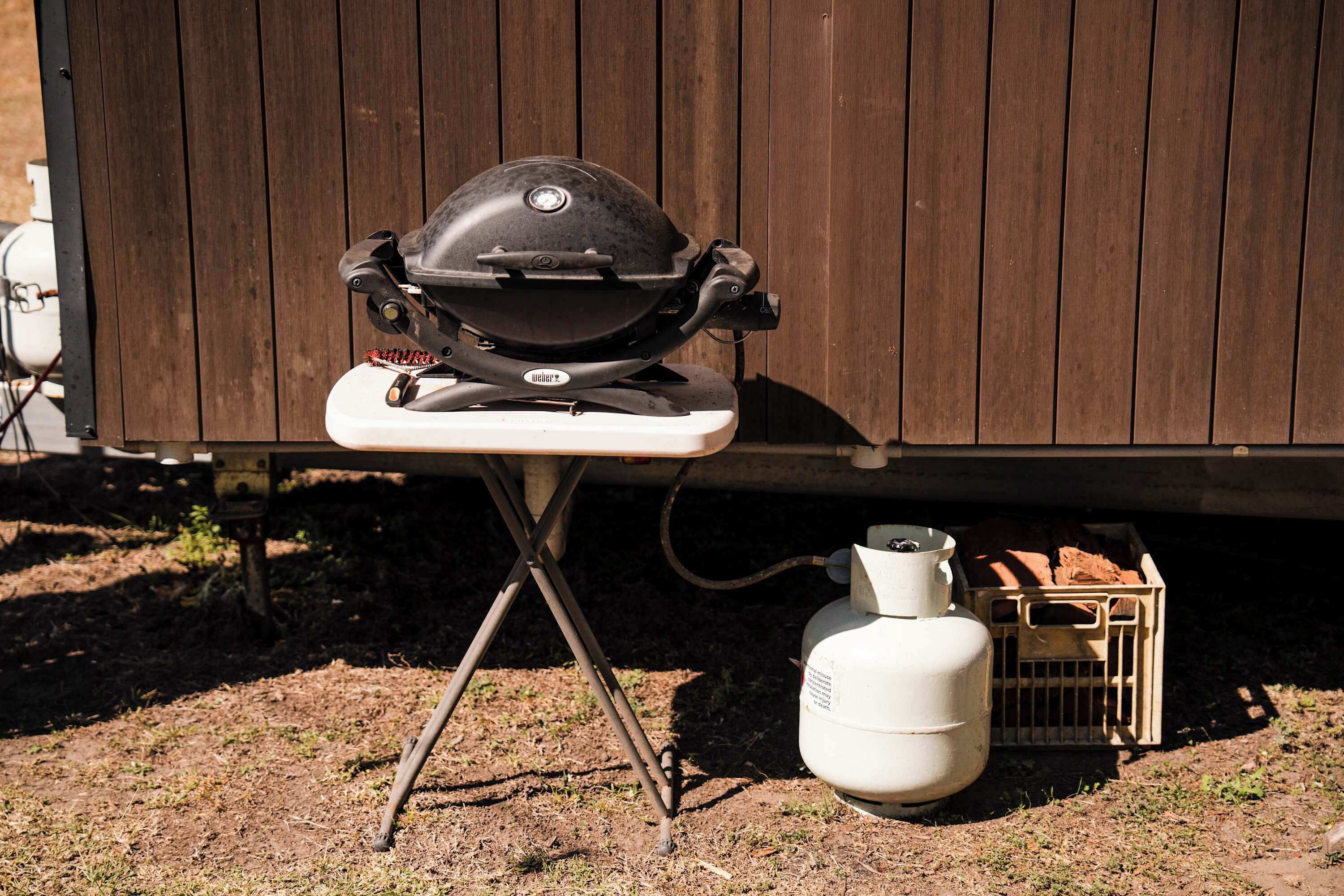 A portable Weber grill on a foldable stand, a white propane tank, and a wooden crate with firewood.