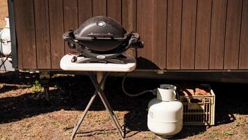 A portable Weber grill on a foldable stand, a white propane tank, and a wooden crate with firewood.