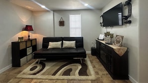 Un salon avec un canapé noir, un tapis à motifs, une table d’appoint avec une lampe, un téléviseur fixé au mur et une fenêtre avec des stores.