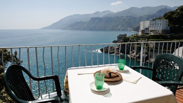 Une table recouverte d’une nappe blanche, deux verres verts et un panier tressé orné d’un coquillage, le tout placé sur un balcon donnant sur la mer et les montagnes.