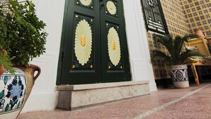 Une grande porte richement décorée, ornée de motifs floraux complexes, flanquée de plantes en pot et d’un mur recouvert de carreaux aux motifs géométriques.