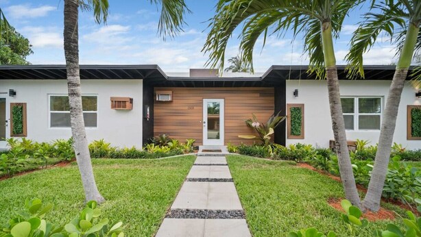 A modern two-story house with a central entrance, surrounded by a well-maintained lawn and palm trees.
