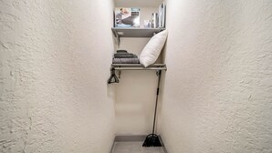 A narrow hallway with a white textured wall, a metal shelf with folded towels and a pillow, and a broom leaning against the wall.