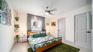 A bedroom with a bed, a ceiling fan, a window with curtains, a door, and a wall-mounted light fixture.