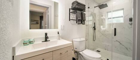 A modern bathroom with a white vanity, a toilet, a glass shower enclosure, and a small window.