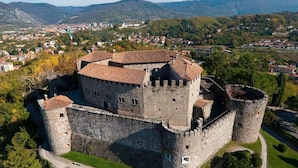 Une vue aérienne d’un château historique en pierre, doté de nombreuses tours et d’un mur d’enceinte, entouré d’une végétation luxuriante et avec un petit village en arrière-plan.