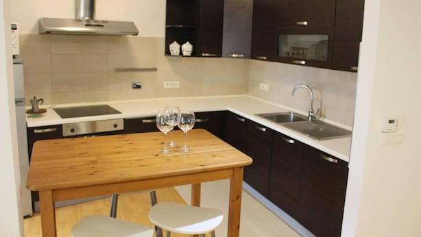Une cuisine compacte avec des meubles de couleur sombre, une hotte de cuisine en acier inoxydable, un évier à deux bacs et une table en bois accompagnée de deux tabourets blancs.