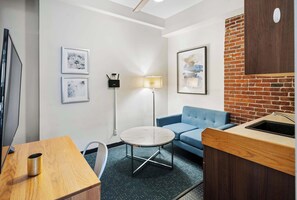 A compact hotel room with a wooden desk, a round table, a blue sofa, a small kitchenette, and two framed artworks on the walls.