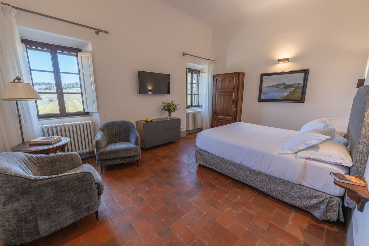 Un dormitorio con una cama, dos sillones, un televisor, un radiador y una ventana con cortinas.