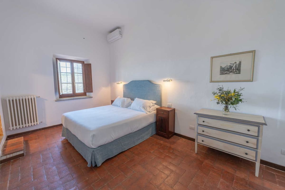 Un dormitorio con cama, mesita de noche, cómoda, radiador, ventana y aire acondicionado de pared.
