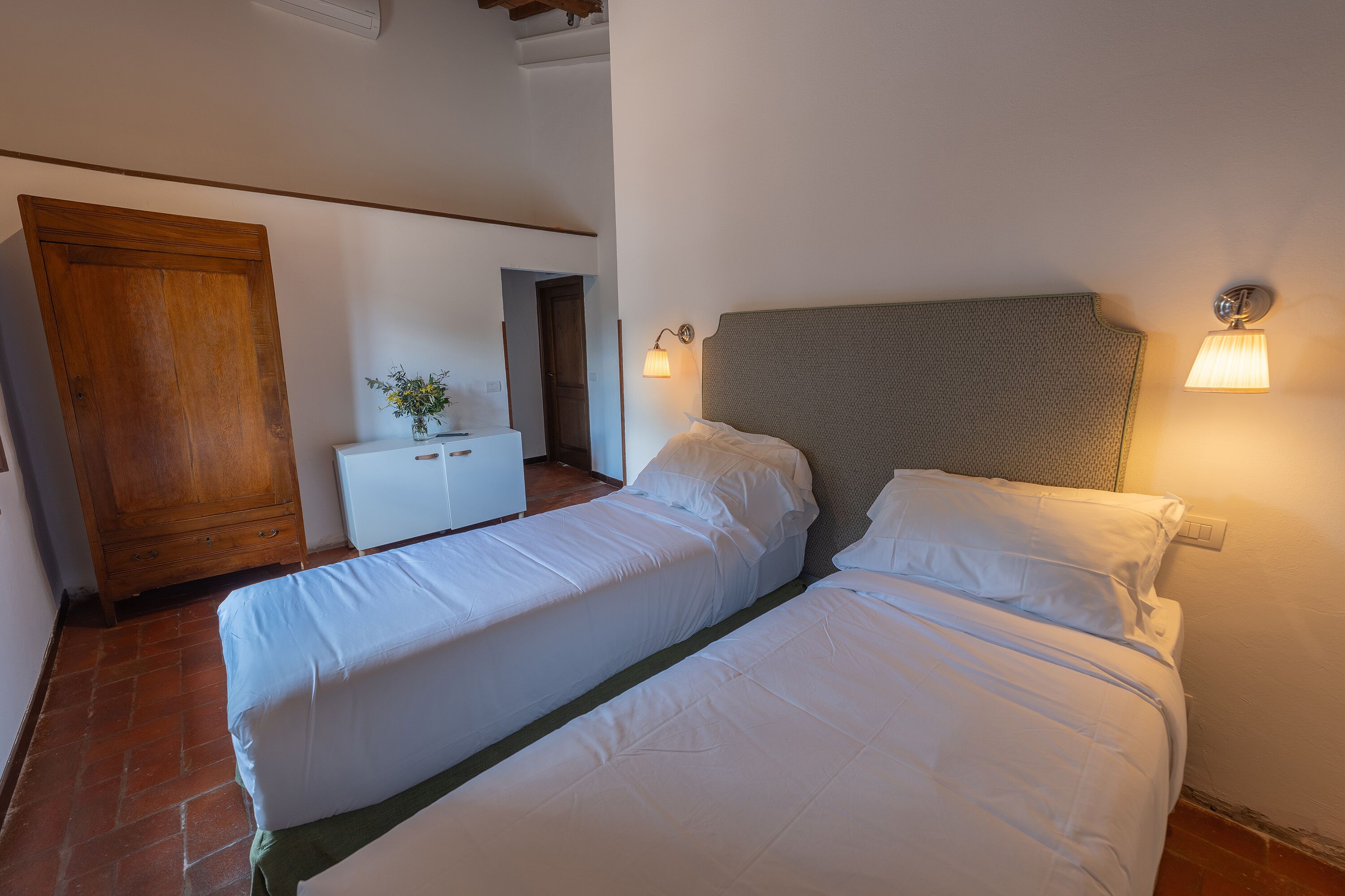 Ein Schlafzimmer mit zwei Betten, einem Holzschrank, einer kleinen weißen Kommode und einer Wandlampe.